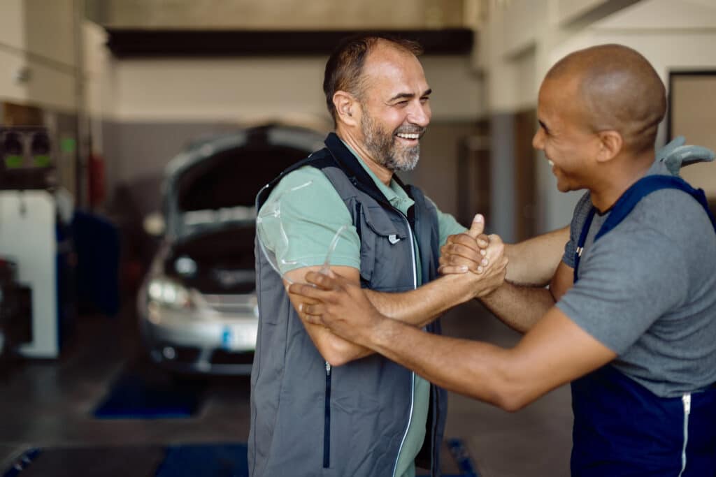 Two happy men shake hands in a bright automotive workshop, showcasing friendship and teamwork.